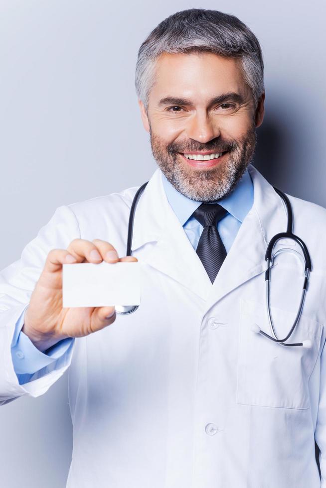 läkare med företag kort. självsäker mogna läkare som visar hans företag kort och leende medan stående mot grå bakgrund foto