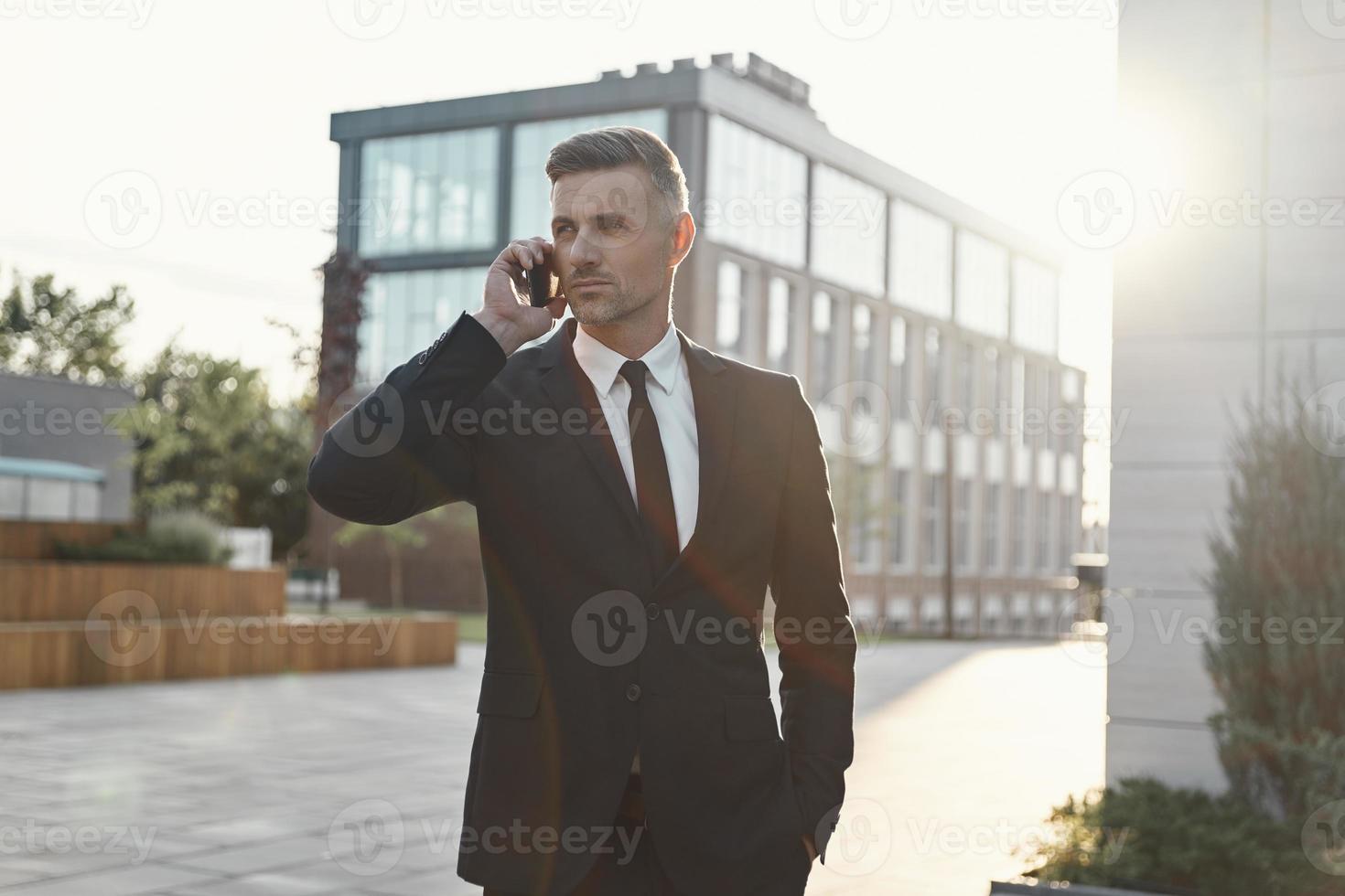 självsäker mogna affärsman talande på mobil telefon medan gående nära kontor byggnad foto