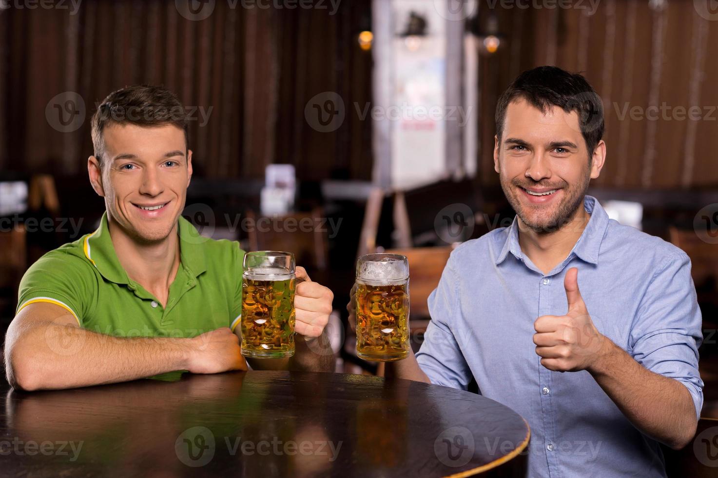 vänner dricka öl. två ung man innehav muggar med öl och leende på kamera foto