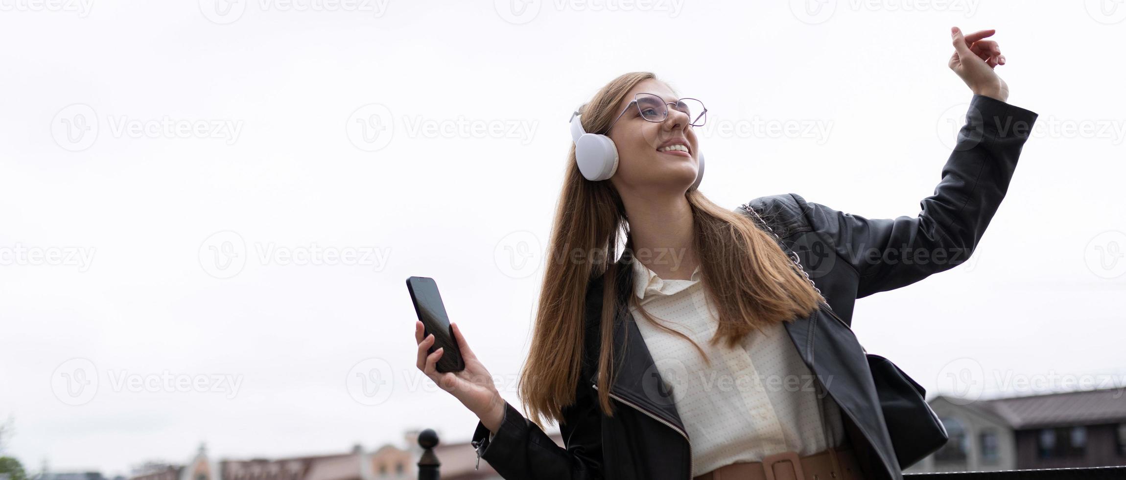 Söt med du en ung kvinna med en mobil telefon i henne händer och hörlurar lyssnar till musik och danser mot de bakgrund av de stad panorama foto