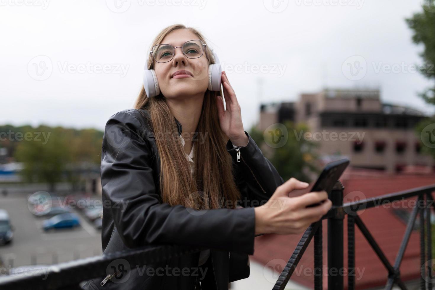 ung kvinna lyssnar med nöje till musik på hörlurar mot de bakgrund av de stad ser eftertänksamt in i de distans foto