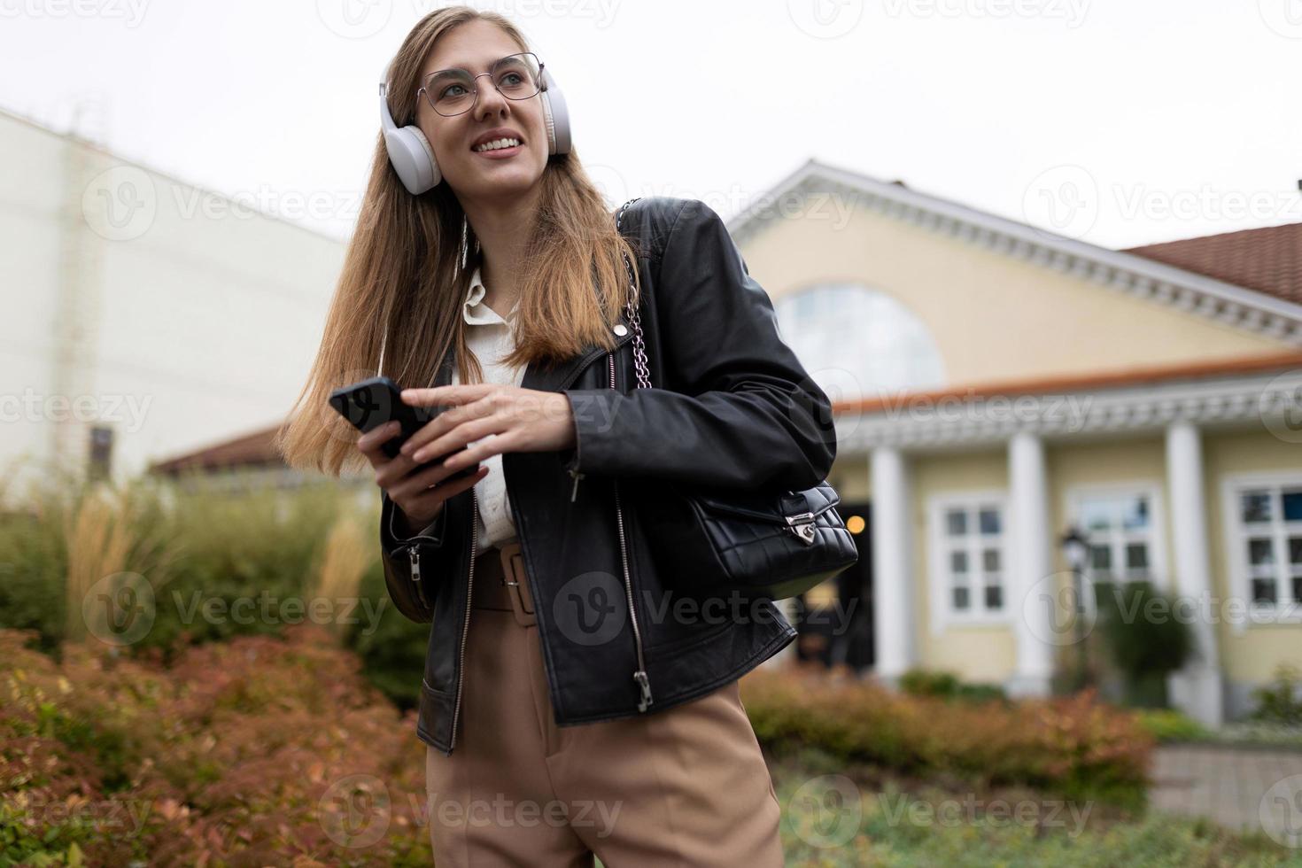 porträtt av en stark ung kvinna studerande i en läder svart jacka och hörlurar med en mobil telefon i henne händer foto