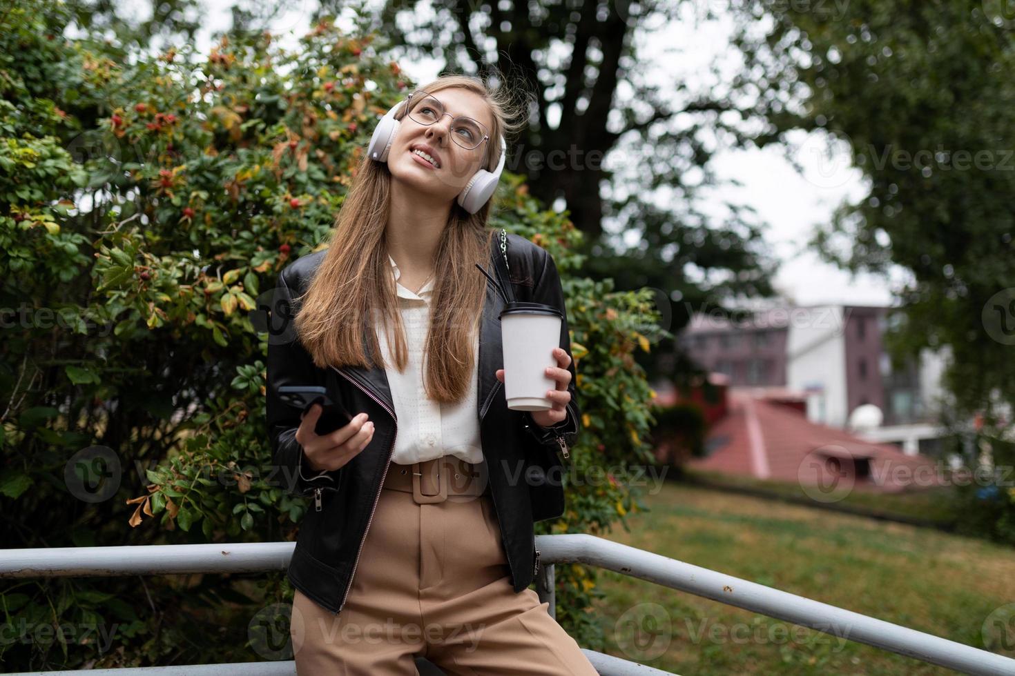 ung kvinna med en kopp av kaffe förbi mobil telefon lyssnande till musik på hörlurar medan avkopplande i de parkera foto