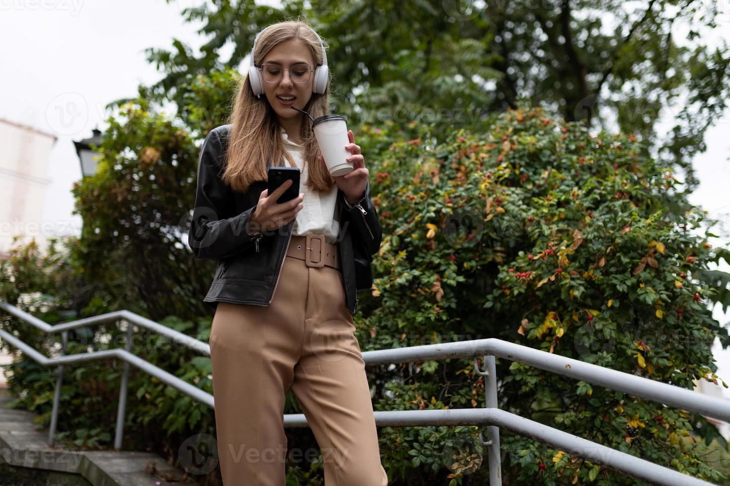 ung kvinna i de stad med en glas av kaffe i hörlurar och en mobil telefon foto