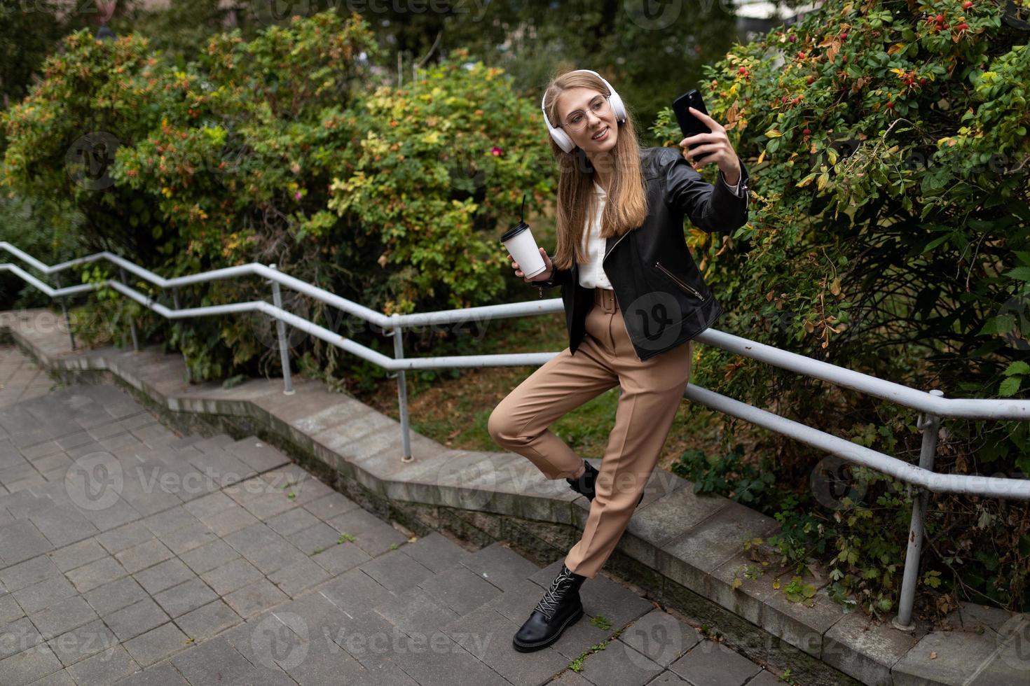 ung kvinna i de parkera lyssnande till musik i hörlurar med en kopp av kaffe och tar en selfie foto