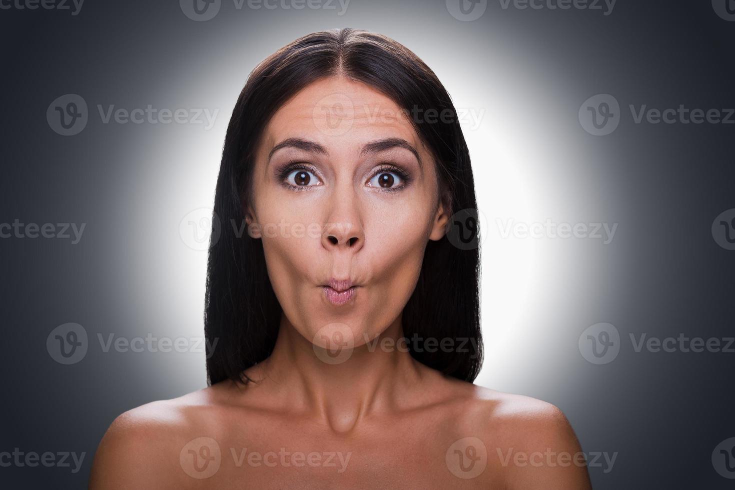 lekfull skönhet. porträtt av skön ung bar överkropp kvinna stirrande på kamera och grimaserande medan stående mot grå bakgrund foto