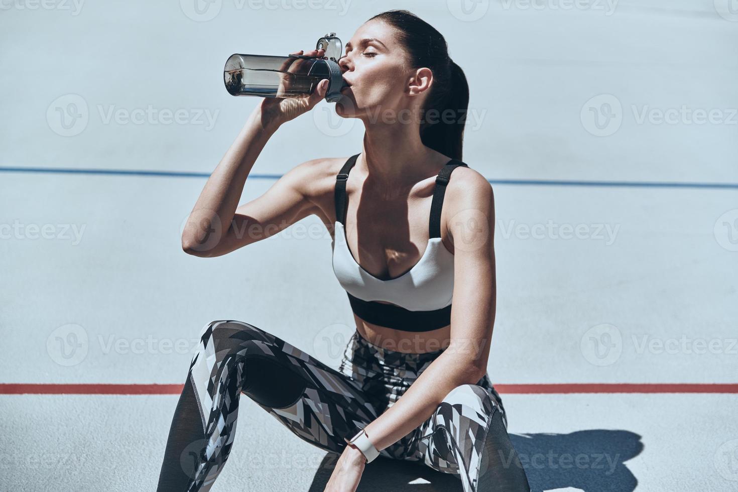 få fräschas upp. topp se av ung kvinna i sporter Kläder dricka vatten medan Sammanträde på de löpning Spår utomhus foto