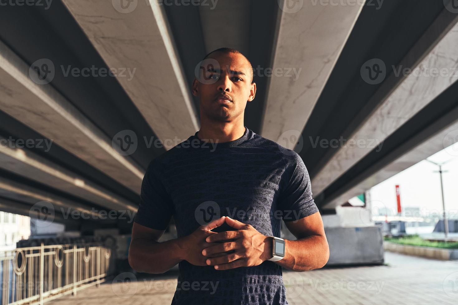 tar tid till resten. stilig ung afrikansk man i sporter Kläder ser på kamera och förvaring händer knäppt medan stående utomhus foto