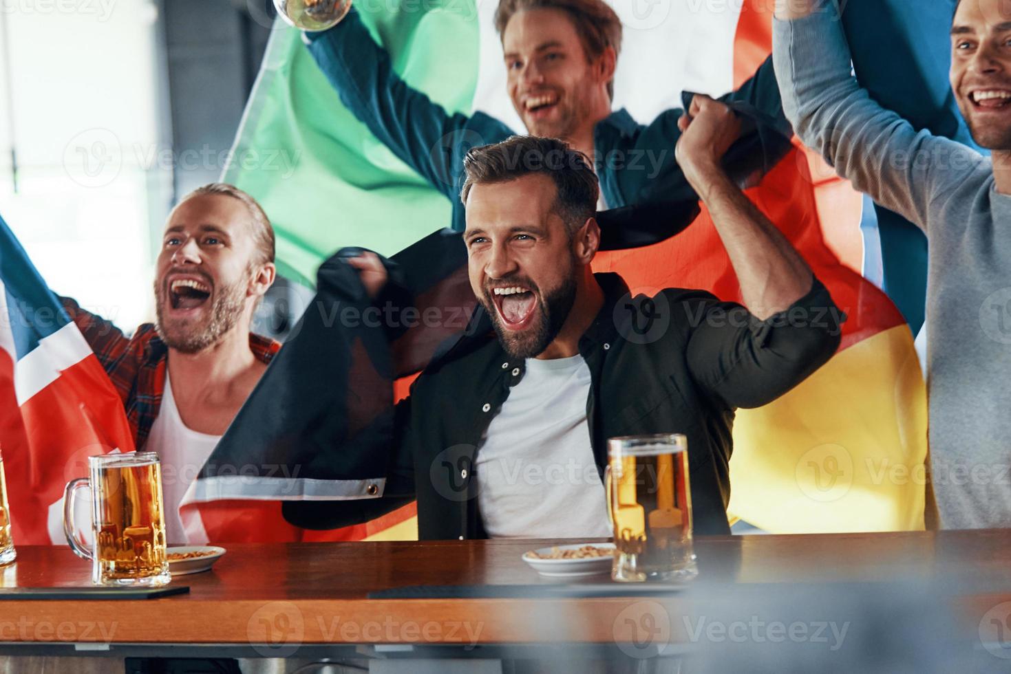 glädjande ung män täckt i internationell flaggor njuter öl medan tittar på sport spel i de pub foto
