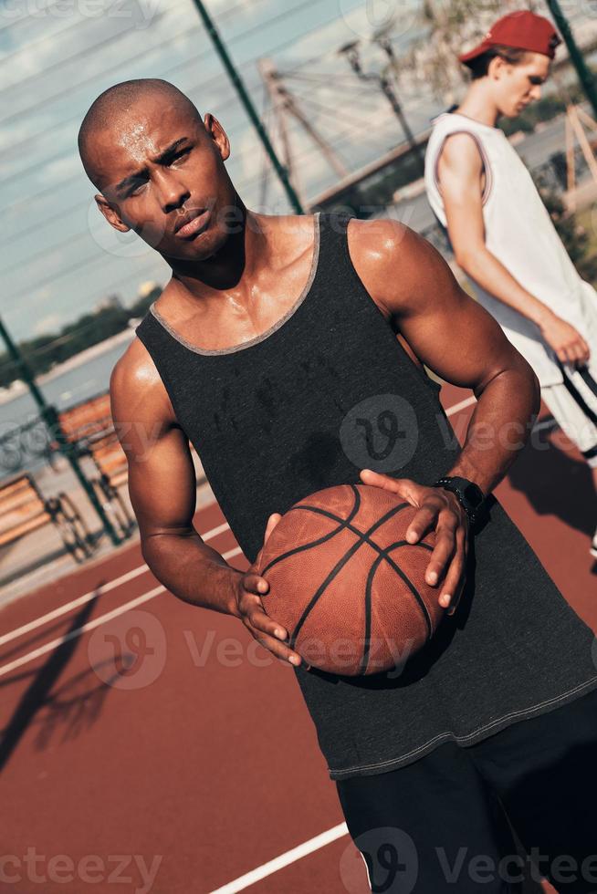 redo till spela. ung afrikansk man i sporter Kläder ser på kamera medan spelar basketboll utomhus foto