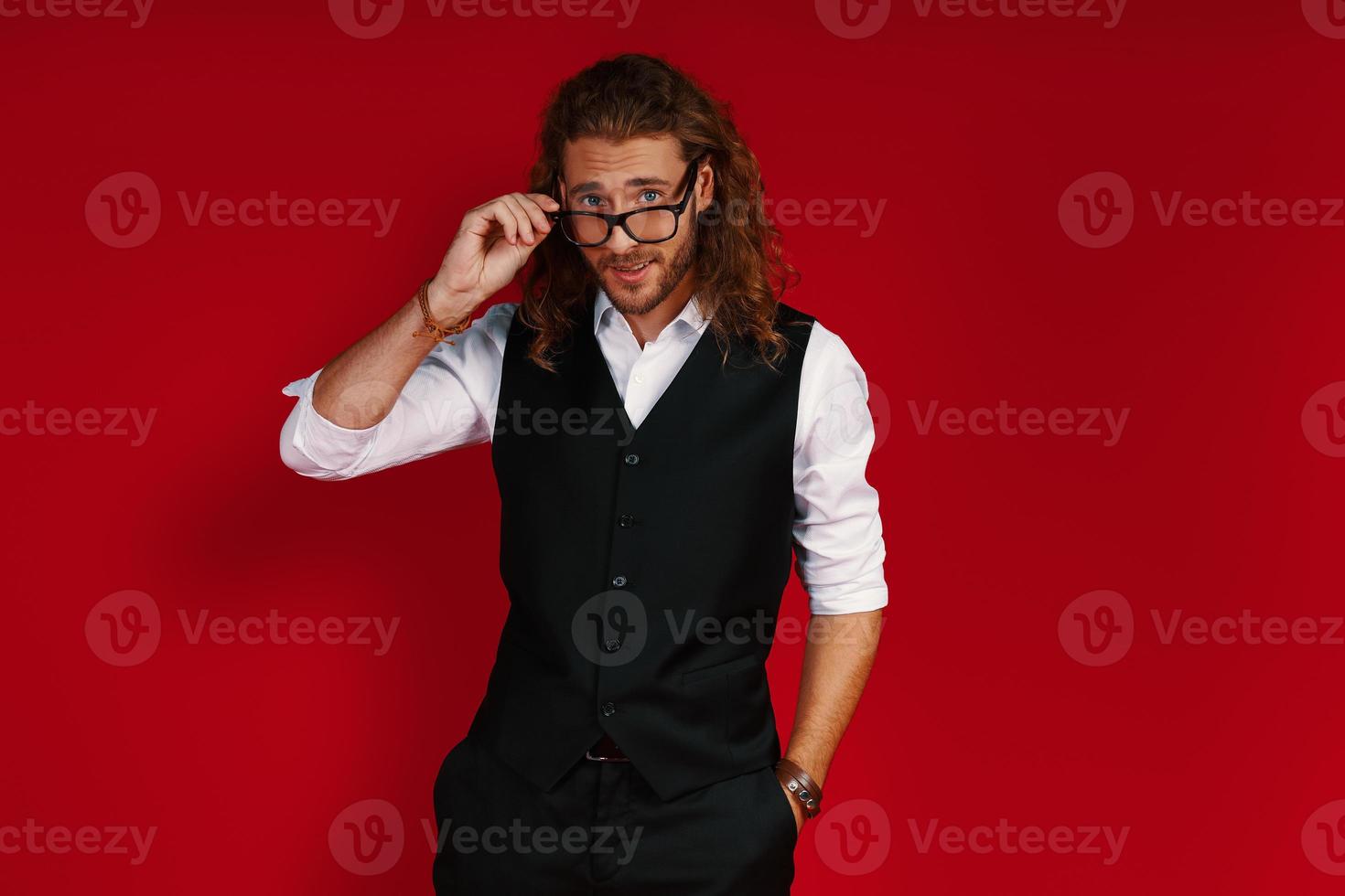 lekfull ung man i kostym ser på kamera med leende och justeras glasögon medan stående mot röd bakgrund foto