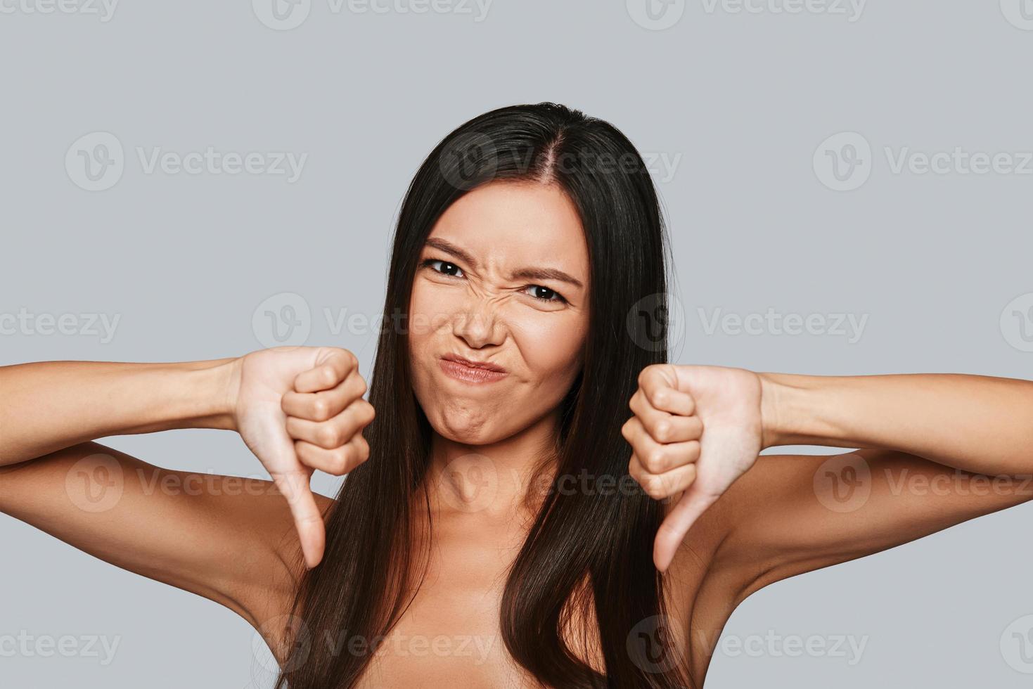 besvikelse. missnöjd ung asiatisk kvinna framställning en ansikte och förvaring tummen ner medan stående mot grå bakgrund foto