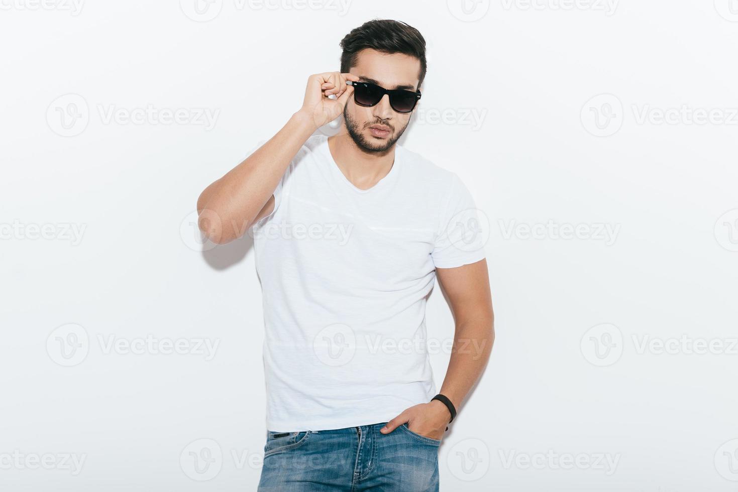 självsäker och stilig. stilig ung indisk man justeras hans solglasögon och ser på kamera medan stående mot vit bakgrund foto