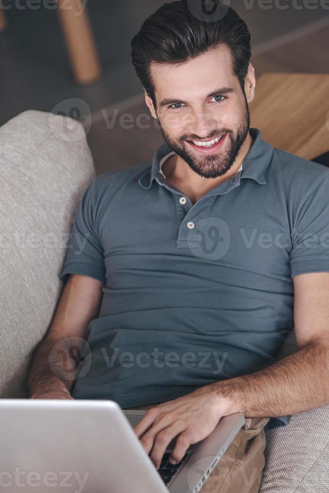 arbetssätt på Hem med nöje. hög vinkel se av stilig ung man använder sig av hans bärbar dator och ser på kamera med leende medan Sammanträde på de soffa på Hem foto