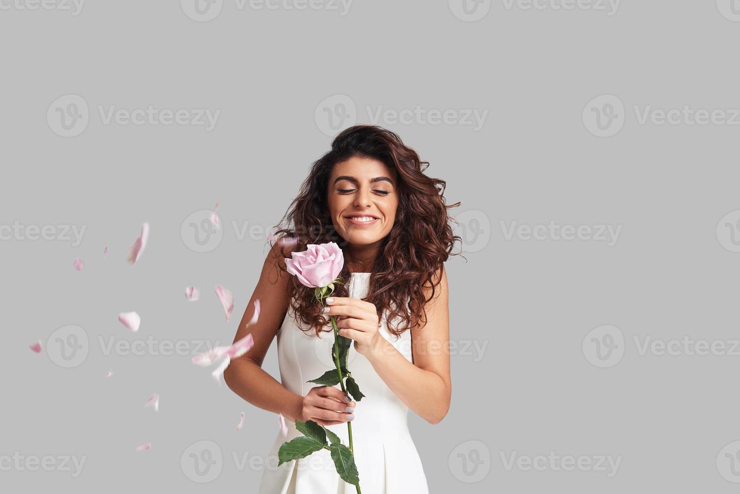 Lycklig ung kvinna leende och innehav en blomma medan stående mot grå bakgrund med reste sig kronblad flygande runt om foto