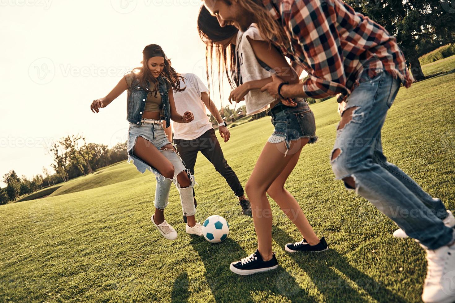 lekfull vänner. grupp av ung leende människor i tillfällig ha på sig njuter trevlig sommar dag medan spelar fotboll utomhus foto