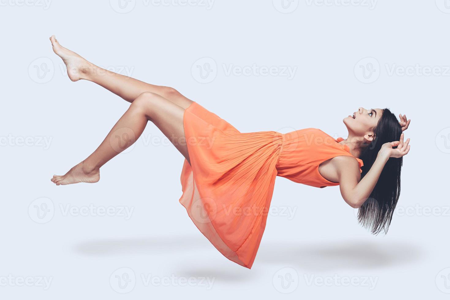 skönhet i i luften. full längd studio skott av attraktiv ung kvinna i orange klänning svävande i luft och ser överraskad foto