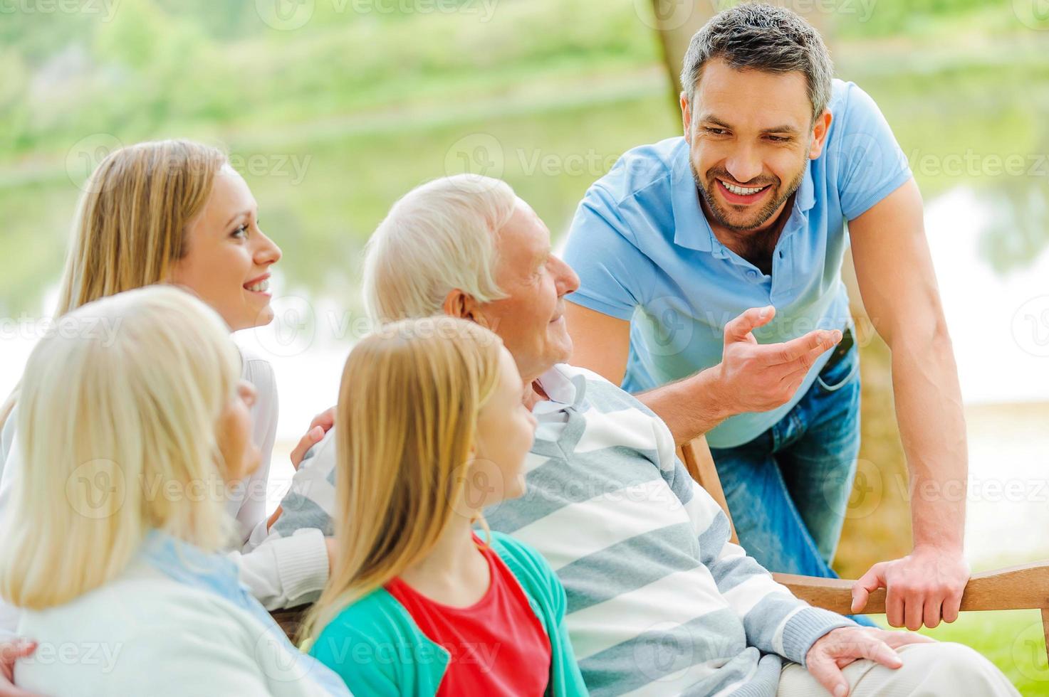 utgifterna tid med familj. Lycklig familj av fem människor talande till varje Övrig och leende medan Sammanträde utomhus tillsammans foto