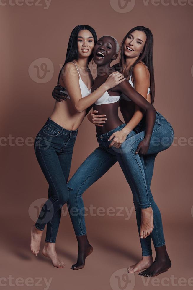 känsla lekfull. full längd av tre attraktiv ung kvinnor ser på kamera och leende medan stående mot brun bakgrund foto
