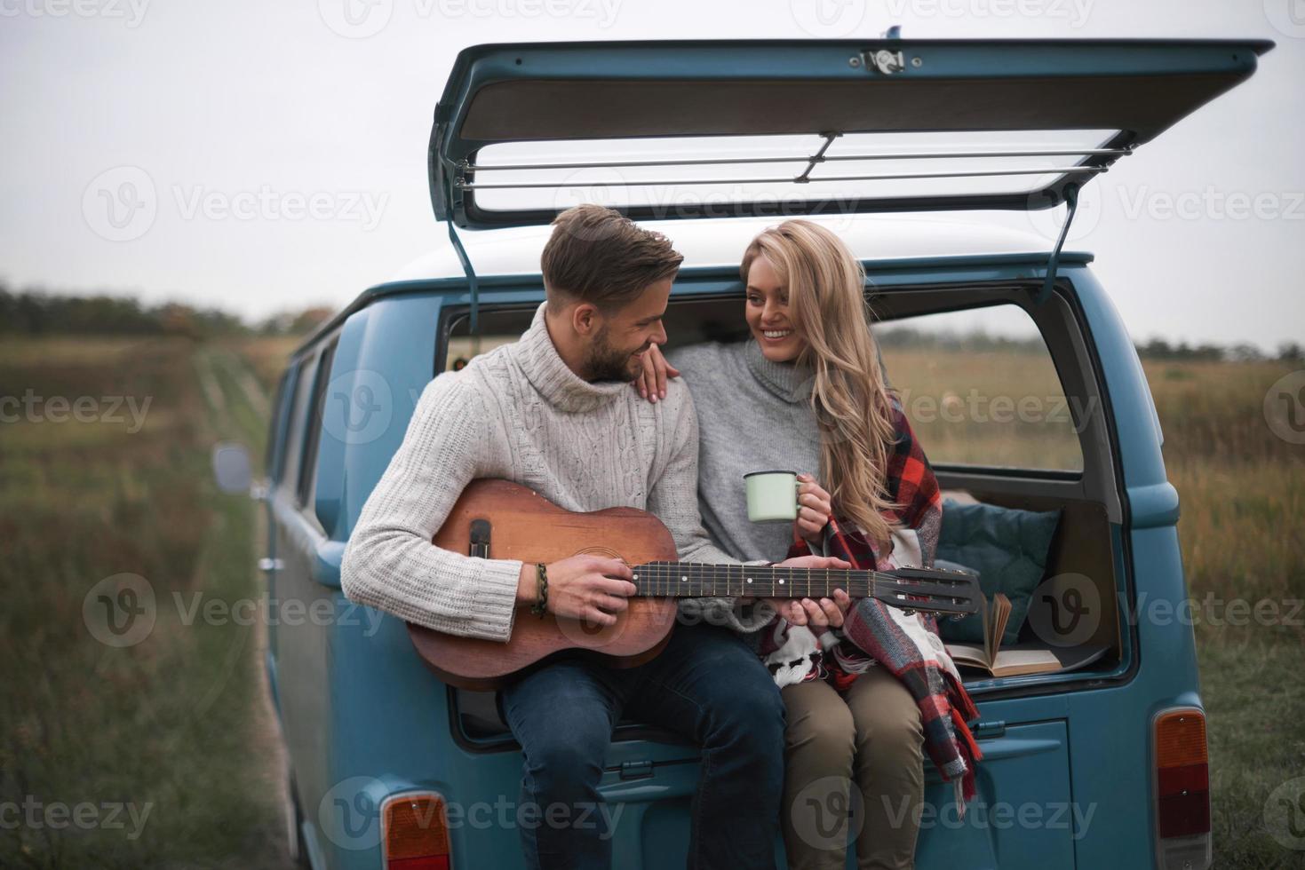 verklig själ kompisar. stilig ung man spelar gitarr för hans skön flickvän medan Sammanträde i de trunk av blå retro stil mini skåpbil foto