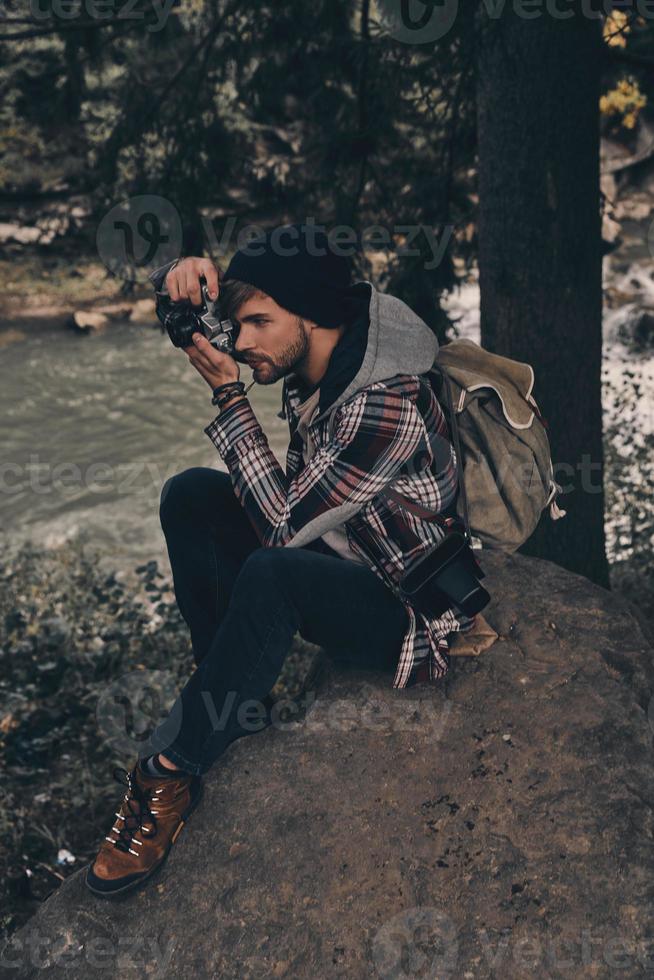 fångande de skönhet. ung modern man med ryggsäck fotografering de se medan Sammanträde i de trän med flod i bakgrund foto