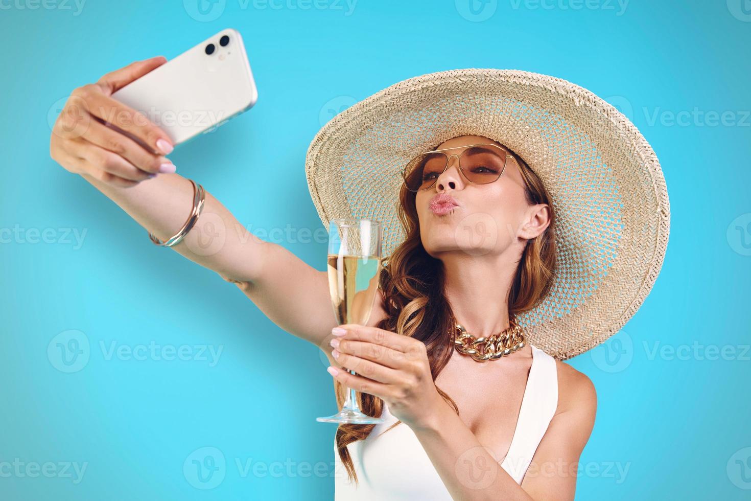 skön ung leende kvinna i elegant hatt innehav champagne flöjt och framställning selfie foto