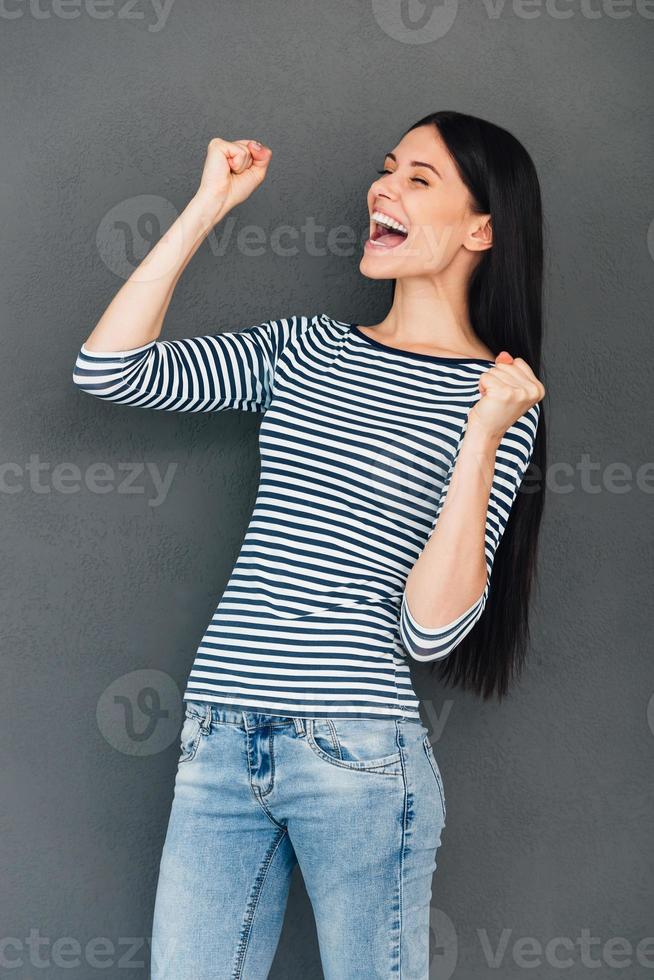 Lycklig vinnare. Lycklig ung kvinna förvaring ögon stängd och gestikulerar medan stående mot grå bakgrund foto