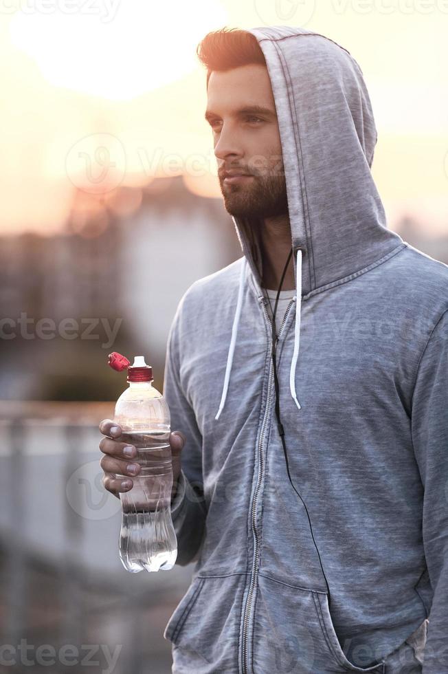 den var Bra träna. självsäker ung man innehav flaska med vatten och ser bort medan stående utomhus foto