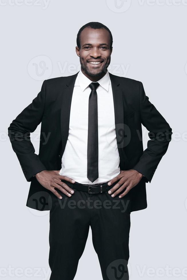 ung och framgångsrik. stilig ung afrikansk man i formell klädsel förvaring händer på höft och ser på kamera medan stående mot grå bakgrund foto