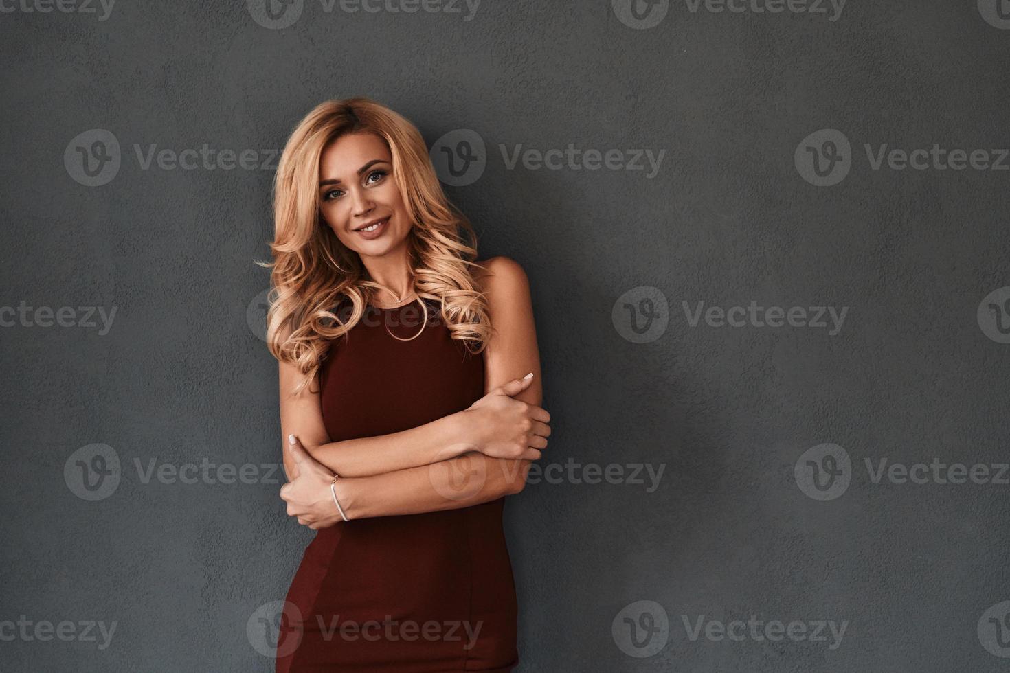 uppriktig leende. attraktiv ung kvinna i klänning förvaring vapen korsade och leende medan stående mot grå bakgrund foto