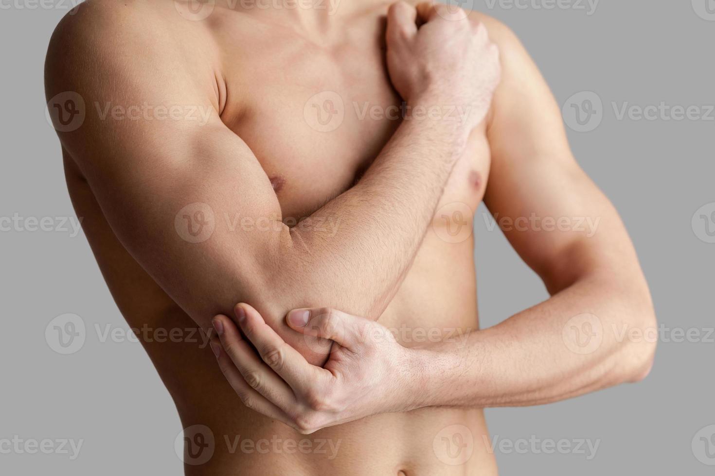 känsla smärta i armbåge. beskurna bild av ung muskulös man rörande hans armbåge medan stående isolerat på grå bakgrund foto