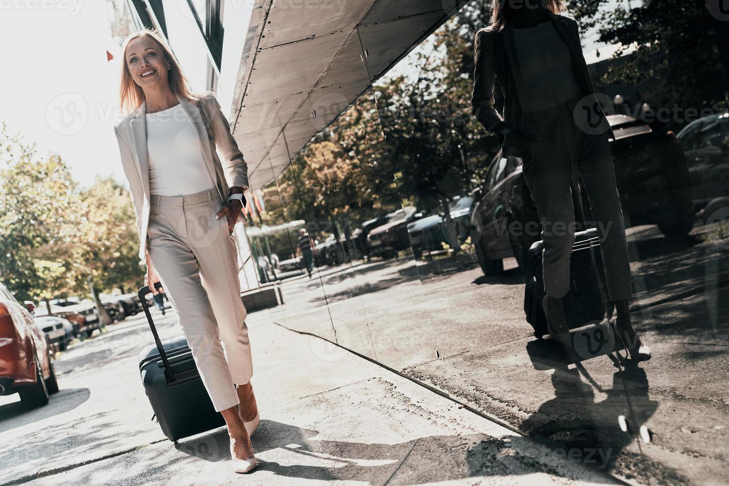 ung företag professionell. full längd av ung kvinna i kostym dragande bagage och leende medan gående utomhus foto