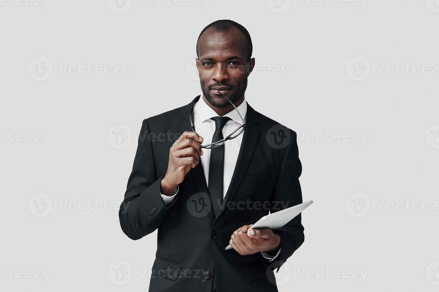 charmig ung afrikansk man i formell klädsel arbetssätt använder sig av digital läsplatta medan stående mot grå bakgrund foto