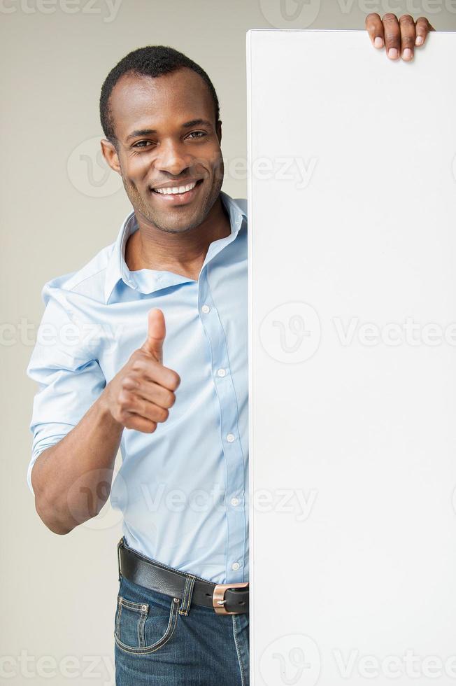 presenter din produkt. glad afrikansk man i blå skjorta lutande på kopia Plats och som visar hans tumme upp medan stående mot grå bakgrund foto