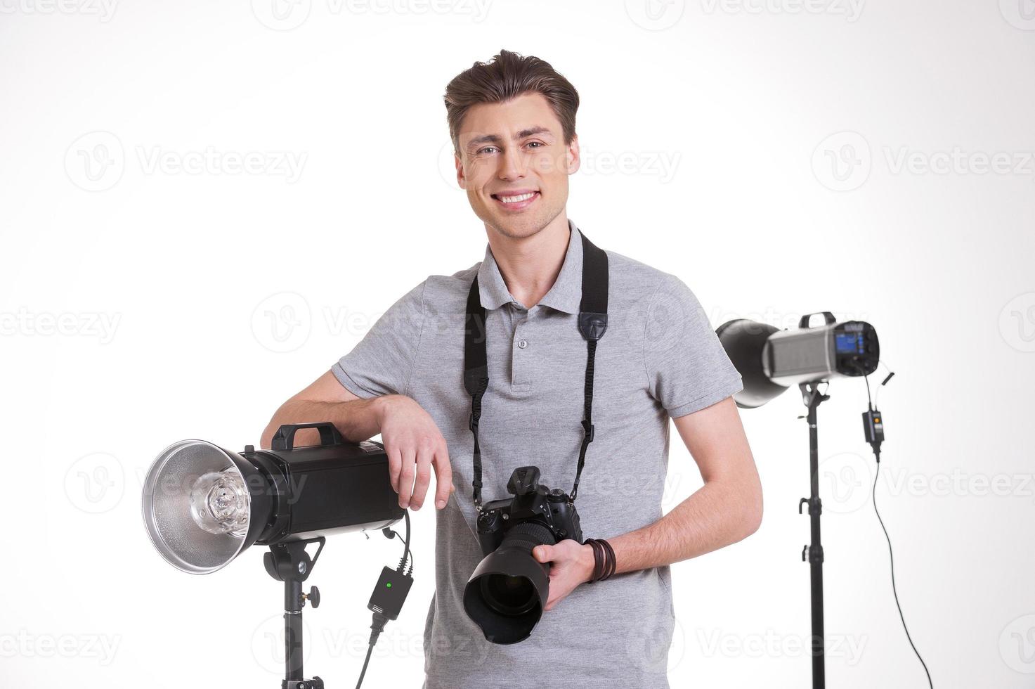 redo för skytte. stilig ung man i polo skjorta innehav digital kamera och leende medan stående i studio med belysning Utrustning på bakgrund foto