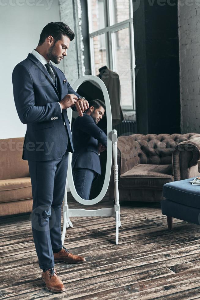 ren elegans. full längd av stilig ung man i full kostym justeras hans jacka medan stående i främre av de spegel inomhus foto