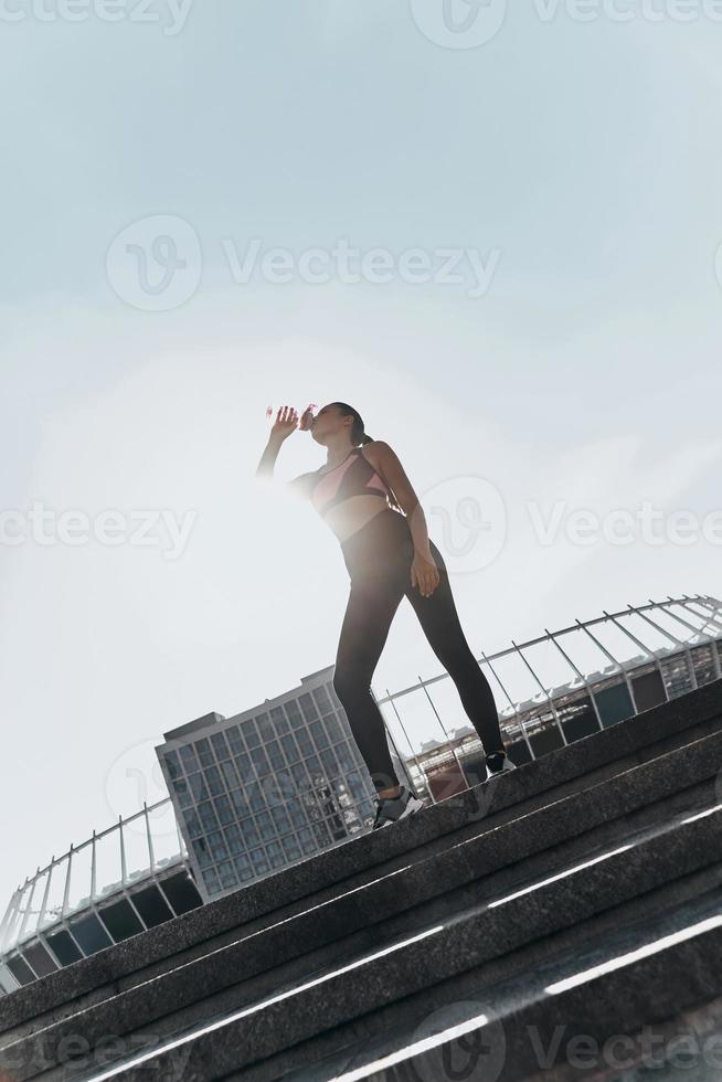 vistas hydrerad. full längd av ung kvinna i sport Kläder dricka vatten medan stående utomhus foto