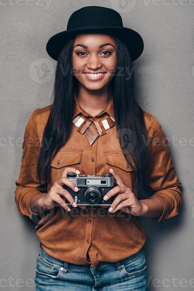 glad fotograf. skön glad ung afrikansk kvinna innehav retro styled kamera och ser på kamera med leende medan stående mot grå bakgrund foto