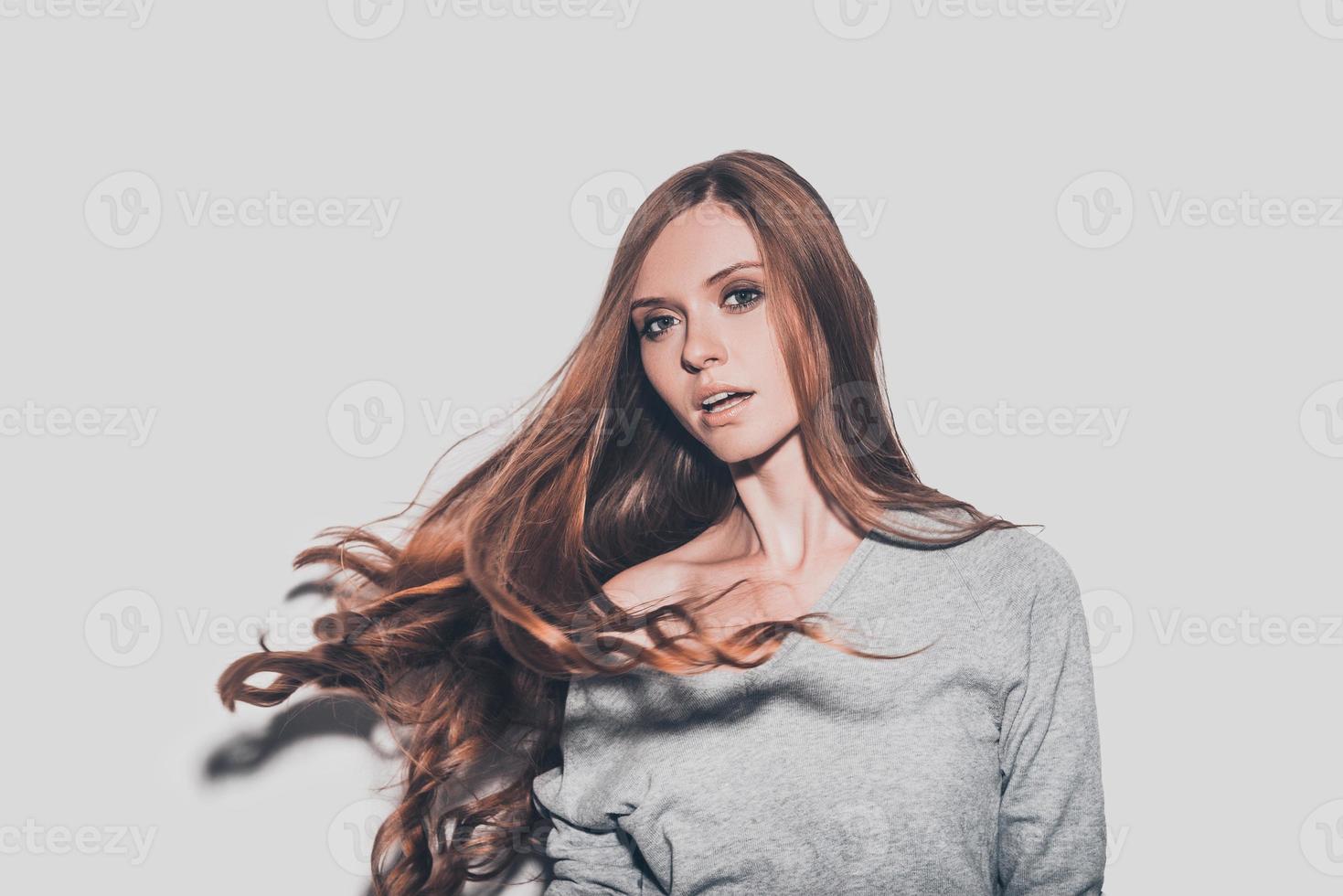 hår i rörelse. porträtt av attraktiv ung kvinna med rufsig hår ser på kamera medan stående mot grå bakgrund foto