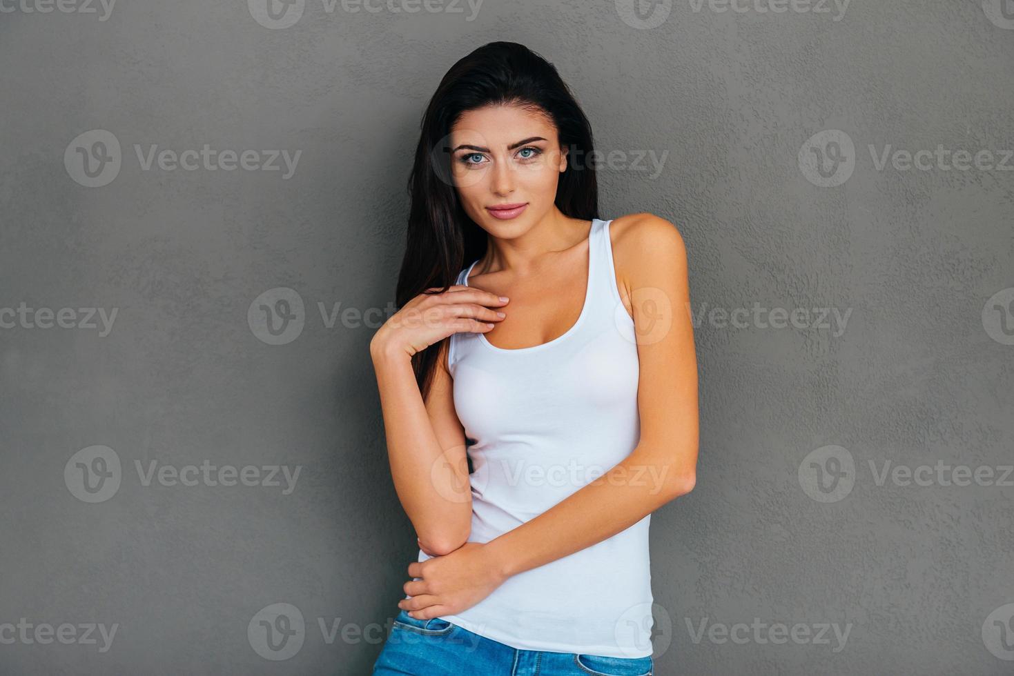 skön förförerska. skön ung kvinna i vit tank topp ser på kamera medan stående mot grå bakgrund foto