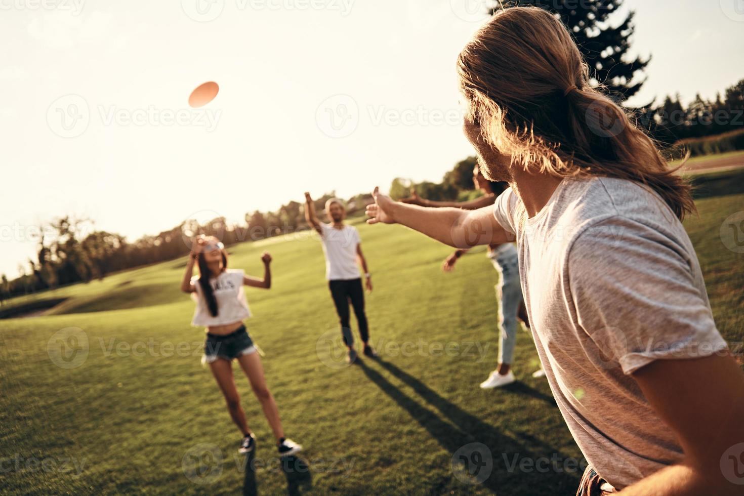 grupp av ung människor i tillfällig ha på sig spelar frisbee medan utgifterna sorglös tid utomhus foto