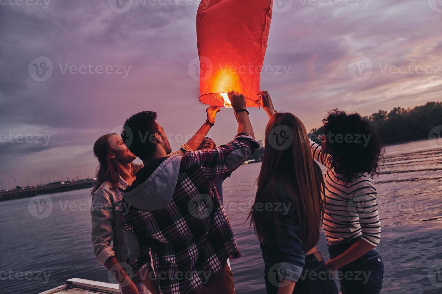 har roligt. grupp av ung människor i tillfällig ha på sig innehav himmel lykta medan stående på de pir foto