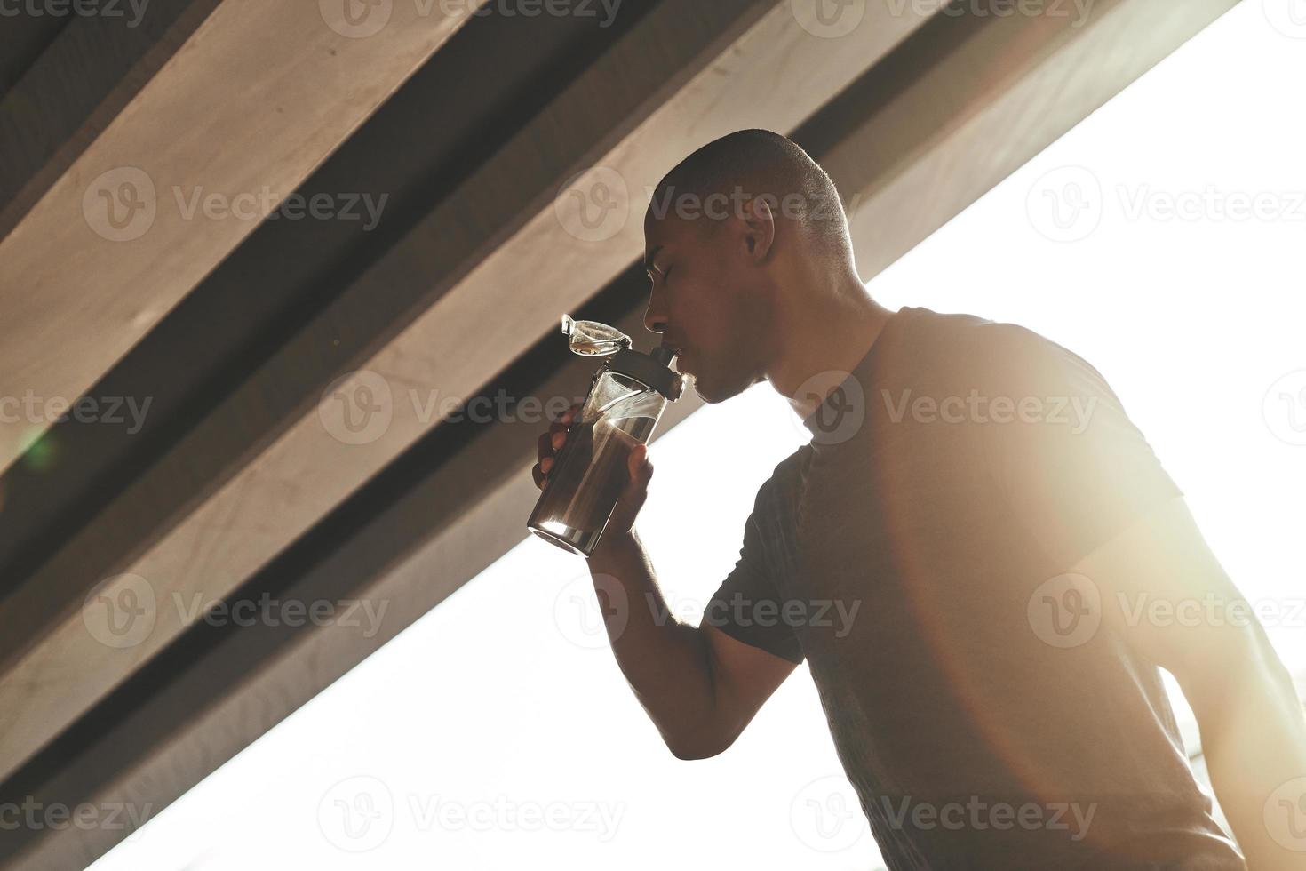 så törstig. ung afrikansk man i sporter Kläder dricka vatten medan stående utanför foto