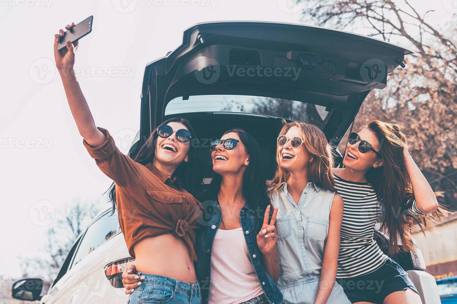 detta är Varför vi ta väg resor fyra skön ung glad kvinnor framställning selfie med leende medan stående nära bil trunk foto