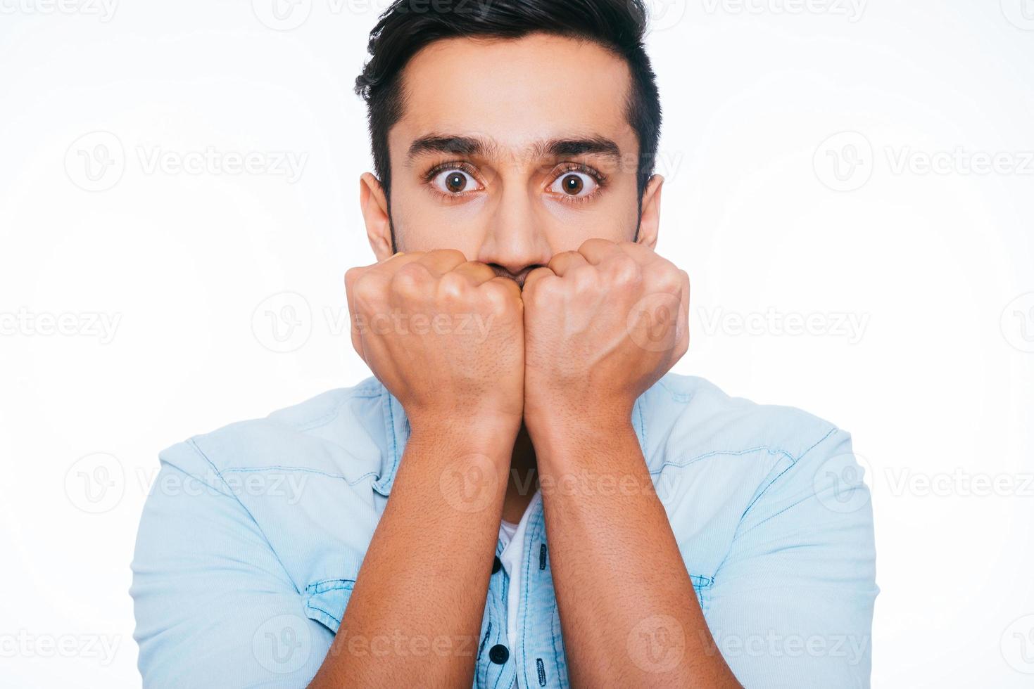 känsla chockade och livrädd. livrädd ung indisk man innehav nävar nära ansikte och stirrande på du medan stående mot vit bakgrund foto