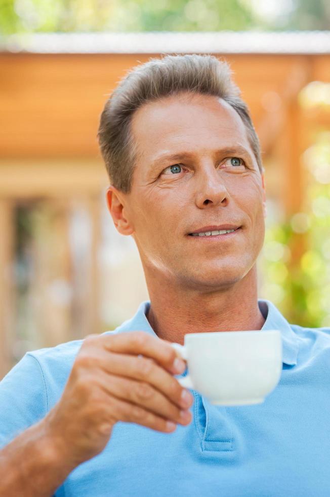 dag drömma med kopp av kaffe. glad mogna man dricka kaffe och leende medan Sammanträde utomhus med hus i de bakgrund foto