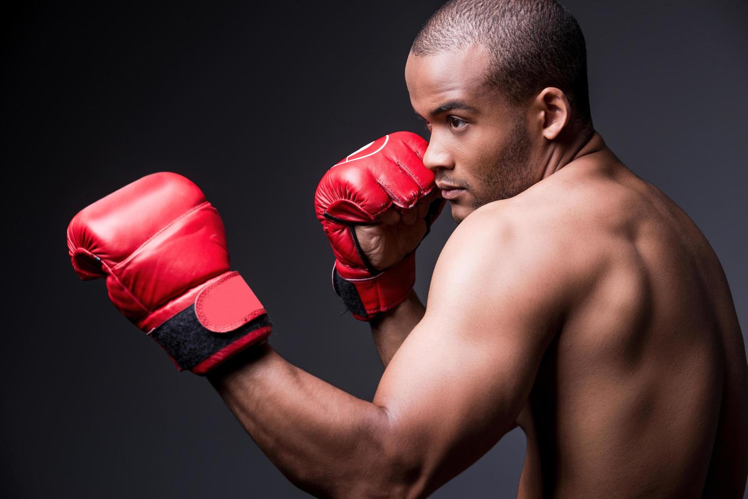 Träning hans boxning Kompetens. sida se av ung bar överkropp afrikansk man i boxning handskar utövar medan stående mot grå bakgrund foto