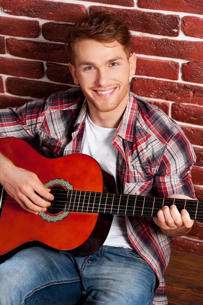 stilig virtuos. topp se av stilig ung man spelar akustisk gitarr och leende medan lutande på de tegel vägg foto