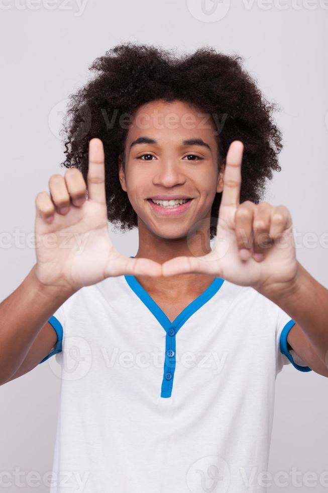 finger ram. glad afrikansk tonåring ser genom en finger ram och leende medan stående isolerat på grå bakgrund foto