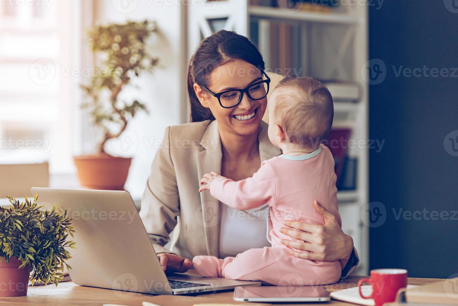 arbetssätt tillsammans är så roligt glad ung skön affärskvinna ser på henne bebis flicka med leende medan Sammanträde på henne arbetssätt plats foto